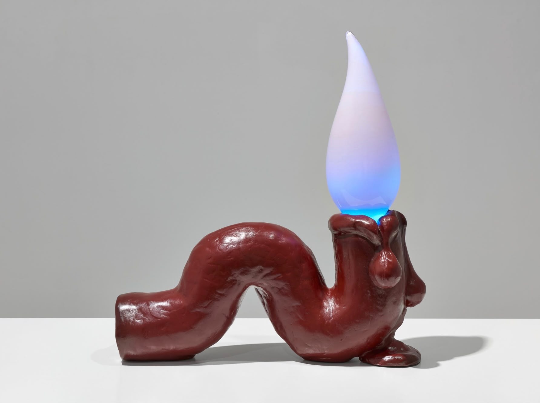 Tai Shani, Haunted by a Million Small Suns (Cardinal), 2025. A low, winding deep-red form like a melted candlestick, with drips at its base, topped by a glowing glass flame shading from blue to lilac.